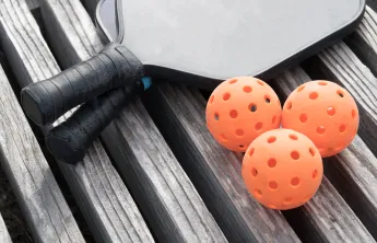 Two pickleball paddles and three pickleballs sitting on a wooden surface.
