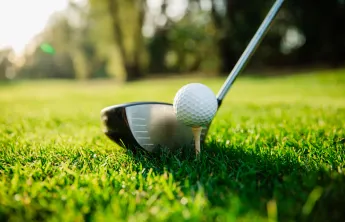 A close-up image of a golf ball on a tee. A golf club is poised near it.