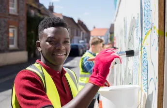 An African American teen wearing a red shirt, gloves, and high-visibility safety vest volunteers outdoors. He is smiling at the camera while painting a wall and holding a white bucket.