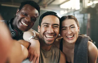 A group of three young adult friends at the gym. They're smiling into the camera.