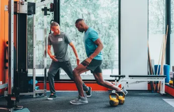 A personal trainer instructs his client in exercise forms and techniques in a gym with orange walls, gray carpet, and large windows. The men are both wearing athletic clothing.