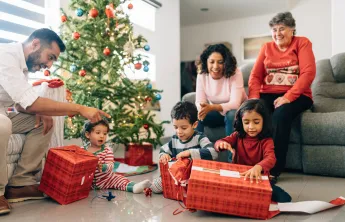 A multigenerational family sitting together on Christmas morning. They are watching three young children open presents.