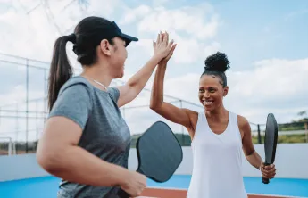 Two women holding Pickleball racquets outdoors. They are smiling and high-fiving each other.