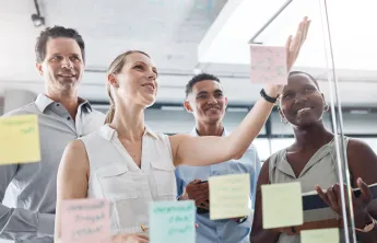 A diverse group working on a vision board together in the form of Post-Its on a glass wall.