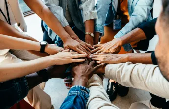 A diverse group of people standing in a circle. They are placing their hands in the center of the group.