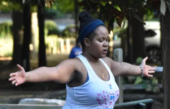 Person practicing yoga outdoors on a mat, balancing in tree pose with arms extended, surrounded by a shaded park setting with trees and seating nearby.