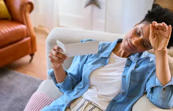 A woman sits on a couch, fanning herself with a card and looking flushed, illustrating a hot flash.