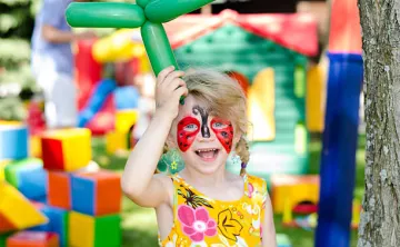 Child with face painting holding a balloon animal in front of bounce house.