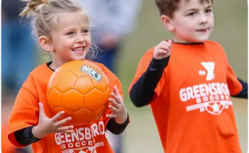 A young boy and girl wearing orange shirts that say Greensboro Soccer in white text. The girl is holding an orange soccer ball and smiling.