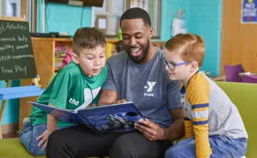 A Y team member reads with two young boys during the YMCA's afterschool program. He is smiling while they look at the open book together.