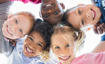 A diverse group of children laughing and playing. They are standing in a circle together and looking down at the camera.