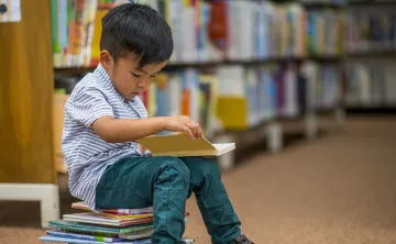 A young boy reading a book. He is sitting on a stack of children's books.