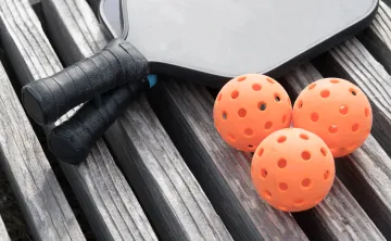 Two pickleball paddles and three pickleballs sitting on a wooden surface.