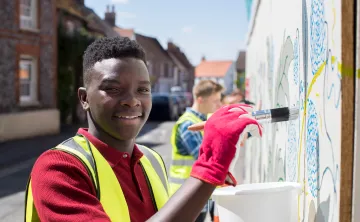 An African American teen wearing a red shirt, gloves, and high-visibility safety vest volunteers outdoors. He is smiling at the camera while painting a wall and holding a white bucket.