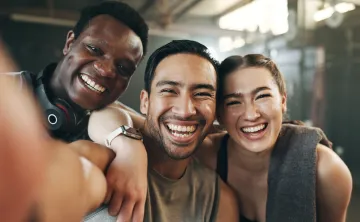 A group of three young adult friends at the gym. They're smiling into the camera.
