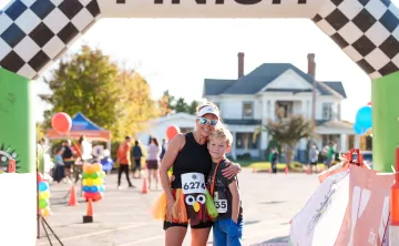 A woman wearing sunglasses and a turkey-themed costume and a young boy stand together outside. They are standing under the road race arch together and smiling at the camera.