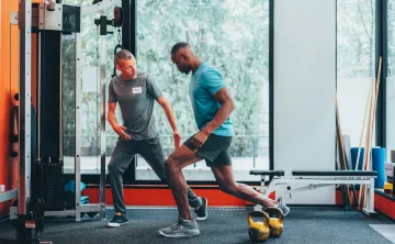 A personal trainer instructs his client in exercise forms and techniques in a gym with orange walls, gray carpet, and large windows. The men are both wearing athletic clothing.