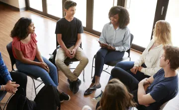 A diverse group of teens sitting together in chairs arranged in a circle. The group is listening to a young woman speak.