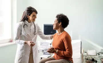 A doctor speaking to a patient in a medical setting. The doctor and patient are both smiling, and the doctor has her hand on her female patient's shoulder.