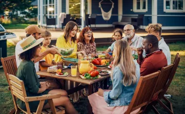 A diverse group of people eating outdoors. They are sitting at a wooden table in wooden folding chairs. A grill is nearby to their left.
