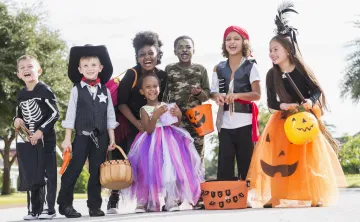 A diverse group of children wearing Halloween costumes. They are standing outside in a group and holding trick or treating bags and buckets.