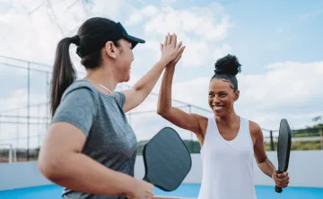 Two women holding Pickleball racquets outdoors. They are smiling and high-fiving each other.
