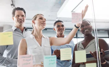 A diverse group working on a vision board together in the form of Post-Its on a glass wall.