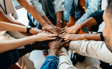 A diverse group of people standing in a circle. They are placing their hands in the center of the group.