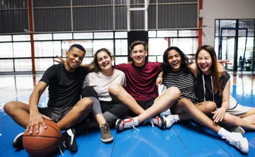 A diverse group of teenagers sitting together on a padded floor mat in a gym. On the left side of the group, a young teen has his hand on a basketball in front of him.