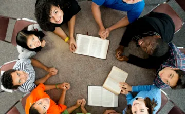 Overhead view of a diverse group of people sitting around the table, with open bibles in the center looking up at the camera smiling.