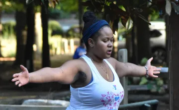 Person practicing yoga outdoors on a mat, balancing in tree pose with arms extended, surrounded by a shaded park setting with trees and seating nearby.
