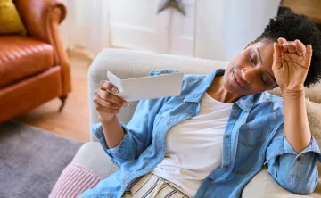 A woman sits on a couch, fanning herself with a card and looking flushed, illustrating a hot flash.