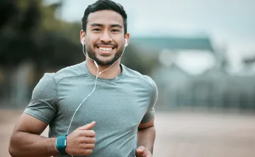 A man running outdoors while wearing earbuds. He is smiling at the camera.