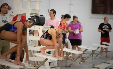 Two swimmers wearing swim caps and goggles prepare to race from starting blocks at an indoor pool during a GCY Makos swim meet.